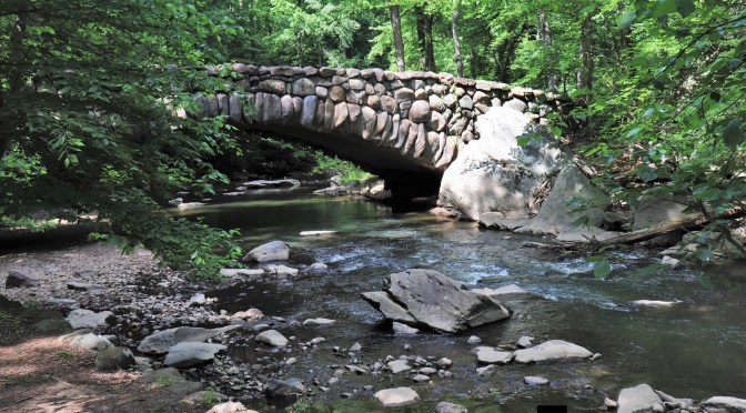 Bach mit Steinen, eine Brücke aus Steinen spannt sich über den Bach, im Hintergrund grüne Bäume