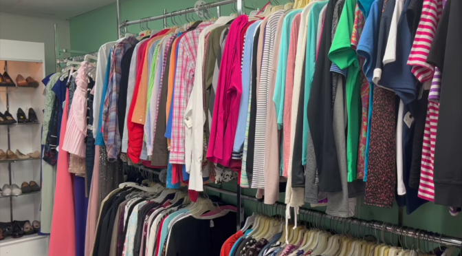Bunte Hemden und Blusen auf einer Kleiderstange, am linken Bildrand ein Regal mit Schuhen