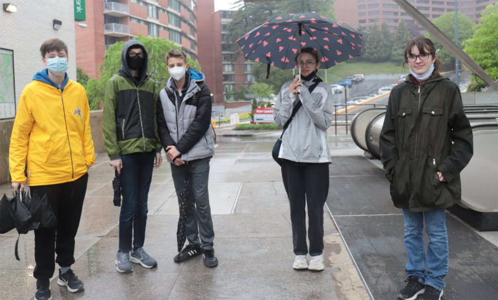 5 Jugendliche in Washington DC vor dem Eingang einer U-Bahnstation. Eine Jugendliche hält einen Regenschirm, die drei Jungen tragen Masken.