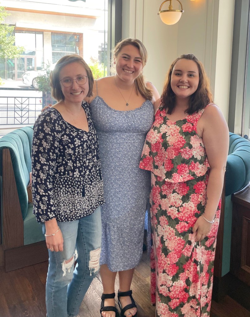 drei lächelnde junge Frauen stehen im Halbkreis vor einem Sofa