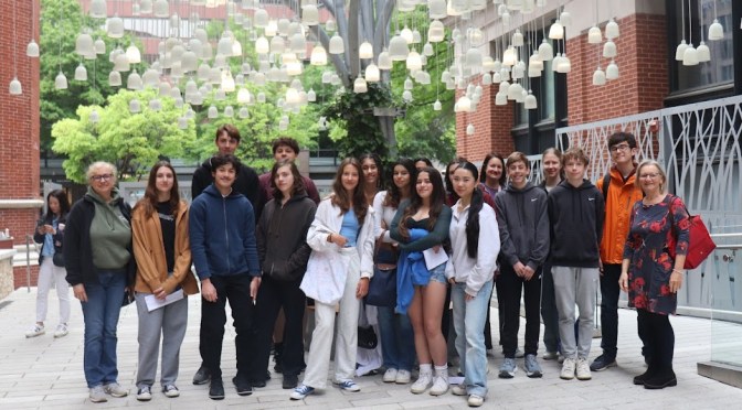 Eine Gruppe junger Leute mit ihren Lehrerinngen auf einem gepflasterten Platz unter hängenden Lautsprechern, die wie kleine weiße Glocken aussehen.