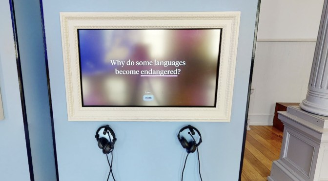 Ein Monitor an einer hellblauen Wand, zwei Kopfhörer hängen darunter. Auf dem Monitor steht: Why do some languages become endangered?