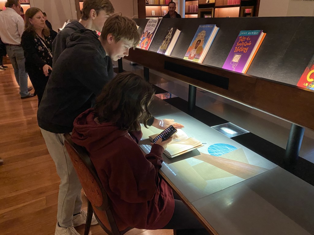 Jugendliche sitzen und stehen vor einem langen Tisch und schauen auf einBuch und ein Handy. Über dem Tisch ist ein Regal mit Büchern zu sehen.